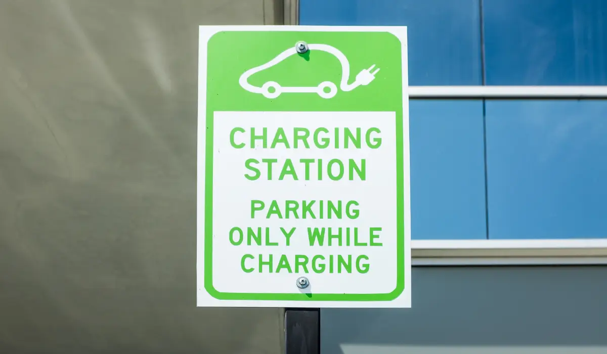 Green and white sign with an electric car icon plugged into a cord, reading “Charging Station: Parking Only While Charging,” mounted on a post in front of a building.